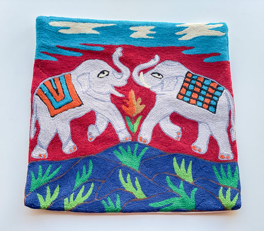 Close up cushion cover with the design of 2 elephants raising their trunks on a red background standing on blue and green grassy hill underneath the blue sky with clouds