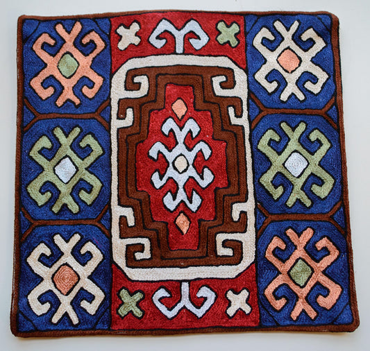 A close up cushion cover depicting a tribal pattern. This is an art silk cushion cover which is hand-embroidered by the artisans of Kashmir, India. The colors of the cushion are dark brown, red, orange, white, light blue and dark blue.
