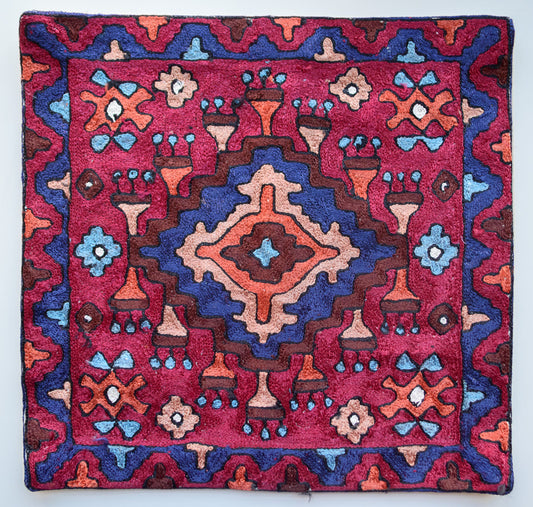 A close up cushion cover depicting a tribal pattern. This is an art silk cushion cover which is hand-embroidered by the artisans of Kashmir, India. The colors of the cushion are dark brown, red, orange, light blue and dark blue.