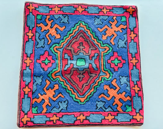 A close up cushion cover depicting a tribal pattern. This is a woolen cushion cover which is hand-embroidered by the artisans of Kashmir, India. The colors of the cushion are light brown, green, red, orange, light blue and dark blue.