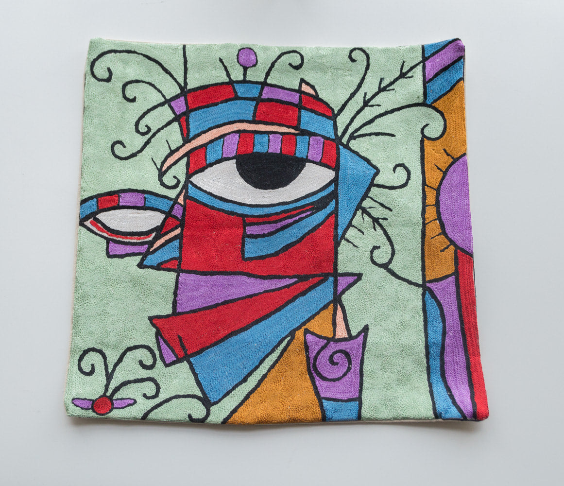 A close up cushion cover depicting modern art form inspired from famous painters like Alfred Gockel and Picasso. The background is light green and the art form shows a funky sideview of a face with one eye. The colors of the face are blue, purple, red, yellow and black and white for the eye. 