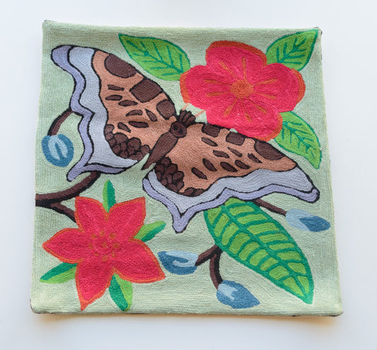A close up cushion cover showing a huge butterfly between 2 flowers. The background color is light green. The flowers are dark pink in color with light and dark green leaves. The butterfly is in the center with colors of light and dark brown, light blue and light purple in it's wings.