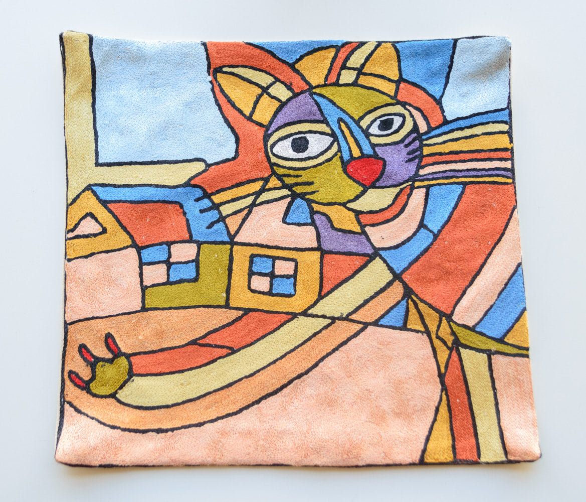 A clsoe up cushion cover showing a geometric cat with her paw stuck out and houses in the background. There are numerous colors of the houses and the cat including blue, purple, red, olive green, light orange, light yellow and dark yellow. The background has the sky top colored in light blue and the ground colored in light orange.