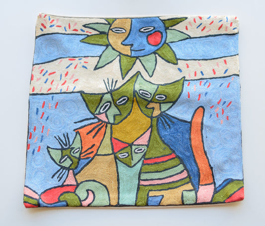 A close up cushion cover having the design of a cat family and a sun face with eyes, nose, smile and a red cheek. The background is mostly light blue with two horizontal cream borders in the background. The main colors of the cats and the sun are green, blue, yellow , red and orange.