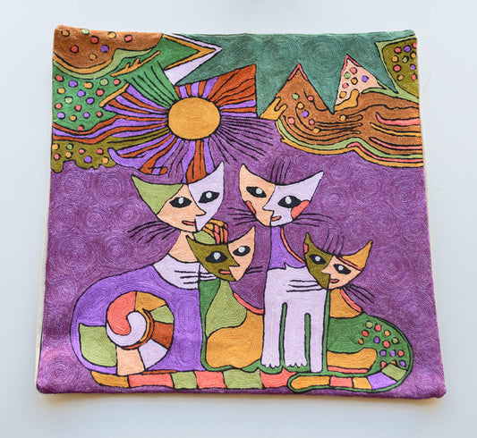 A clsoe up of a cushion cover. The design has a purple background showing the sun and mountains and a cat family of 4- 2 big cats and two small kittens. The cats are colorful having shades of purple, green, cream, red and yellow. It is a very vivid design.