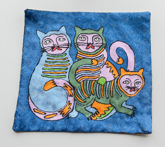 A close up cushion cover showing two big cats with whiskers and a small cat as their child on a blue background. The colors of these cats are light blue, green, light pinkand yellow.