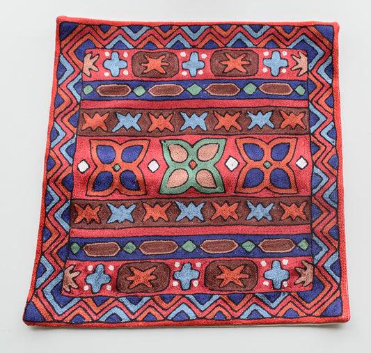 A close up cushion cover depicting a tribal pattern. This is an art silk cushion cover which is hand-embroidered by the artisans of Kashmir, India. The colors of the cushion are light brown, green, red, orange, light blue and purple