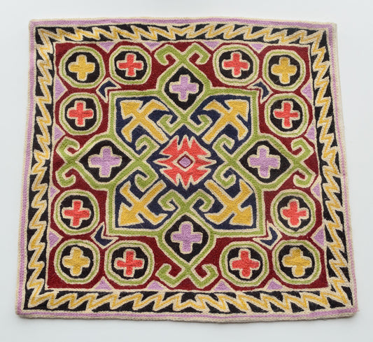 A close up cushion cover depicting a tribal pattern. This is a woolen cushion cover which is hand-embroidered by the artisans of Kashmir, India. The colors of the cushion are dark brown, orange, yellow, green and light purple.
