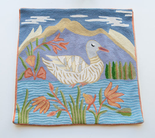A close up cushion cover showing the design of a grey duck in water with the backdrop of blue sky, lavender hill and flowers. The sky has clouds. The hill has some pale yellow to depict sand but mostly is lavender in color. The duck has light shades of white, cream, grey and a red beak. The water has waves and is light and dark blue in color. The flowers are orange with light green and dark green leaves.