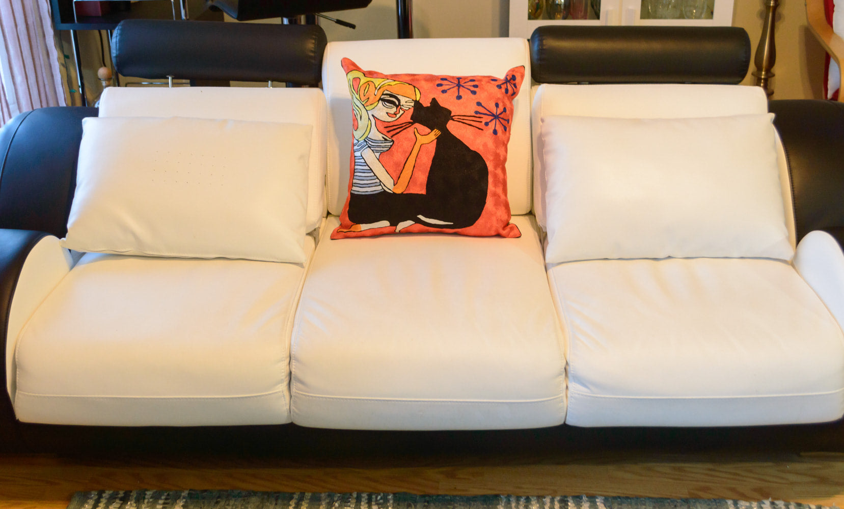 A cushion kept in the center on a white and black sofa. The cushion is having the design of a girl sitting next to her pet black cat and petting her with her hand on her neck. The background is orange color with 3 blue star like shapes.