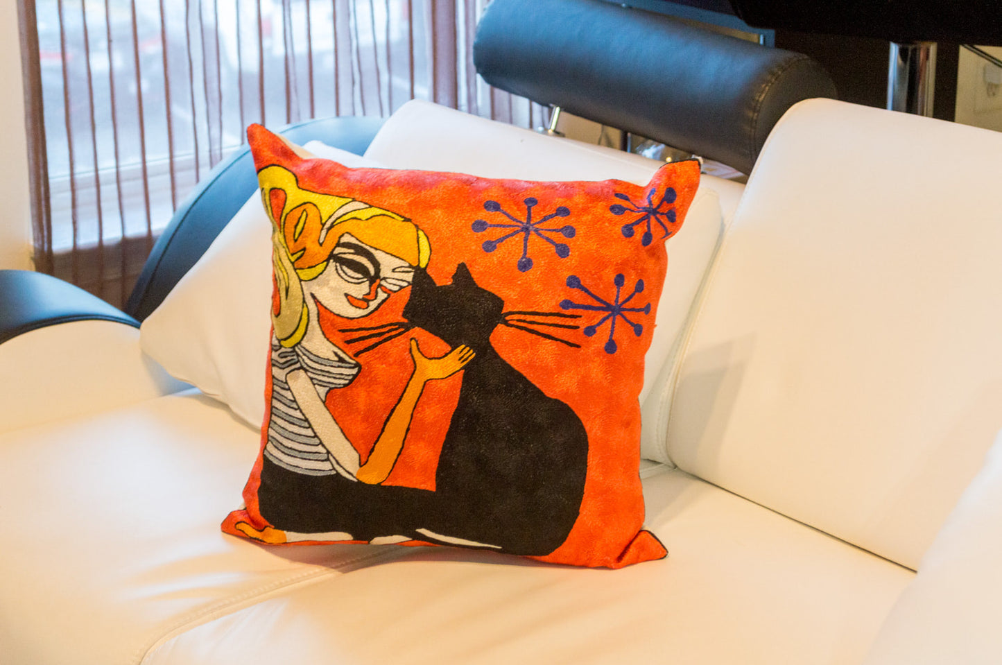 A  cushion kept on a white sofa. The cushion is having the design of a girl sitting next to her pet black cat and petting her with her hand on her neck. The background is orange color with 3 blue star like shapes.