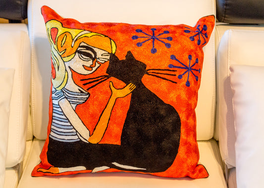 A close up cushion having the design of a girl sitting next to her pet black cat and petting her with her hand on her neck. The background is orange color with 3 blue star like shapes.